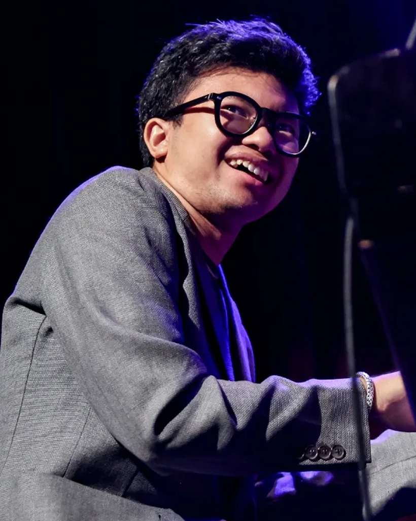Jazz pianist Joey Alexander smiling at the grand piano on stage, wearing round glasses and a grey blazer